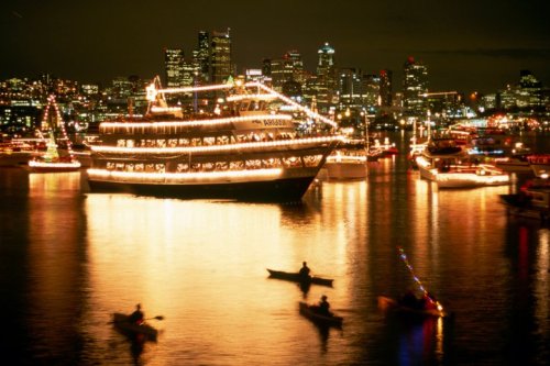 A brightly lit boat surrounded by festive lights, with kayakers on a shimmering waterway and a city skyline in the background.