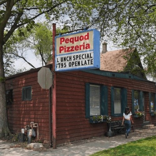 A red wooden building with a sign for Pequod Pizzeria, featuring a lunch special and open late. A person walks by.