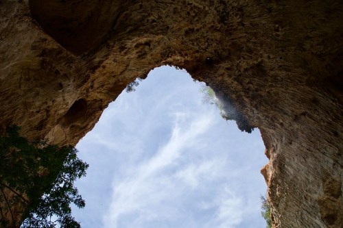 View from below a rocky arch, with a blue sky and wispy clouds visible through the opening. Green foliage at the base.