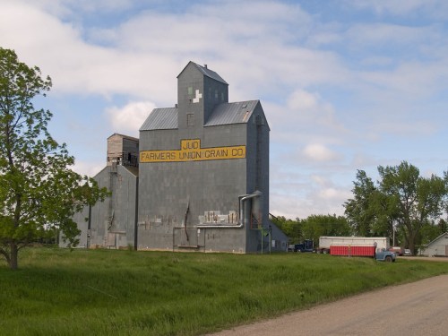 A large gray grain elevator with "Farmers Union Grain Co." painted on it, surrounded by green grass and a dirt road.