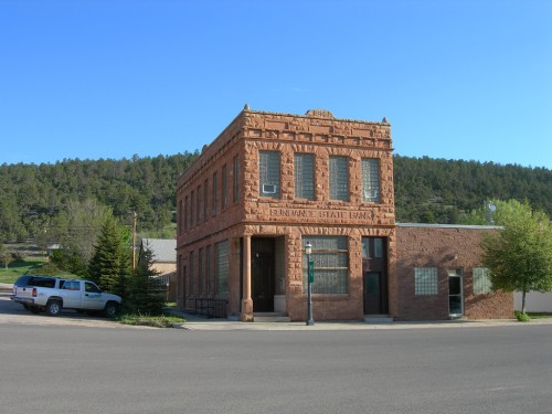 Historic brick building with ornate details, surrounded by trees and parked vehicles in a small town setting.