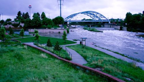 A river flows under a blue arch bridge, surrounded by green parks and trees, with a tower in the background.
