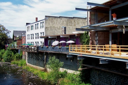 A riverside view of modern buildings with outdoor seating and umbrellas, surrounded by greenery under a blue sky.