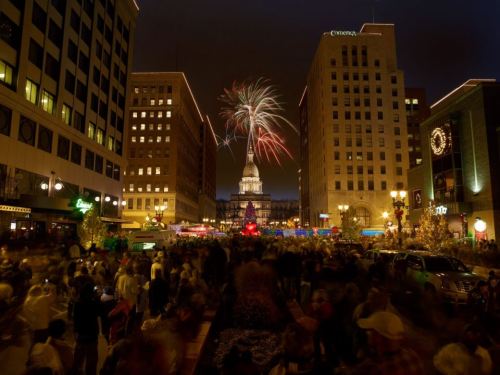 Fireworks illuminate a city street filled with people, with buildings and a clock tower in the background at night.