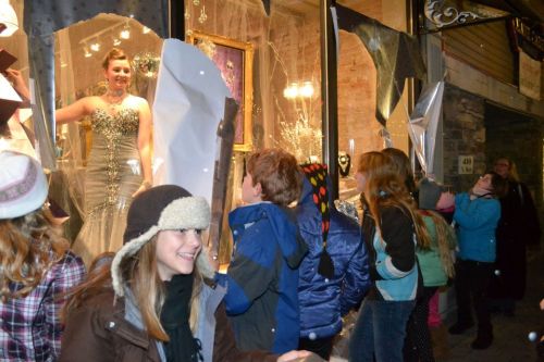 A group of children admire a sparkling dress displayed in a shop window, with festive decorations around them.