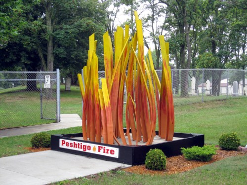 A colorful sculpture resembling flames, titled "Peshtigo Fire," set in a grassy area near a fence and trees.