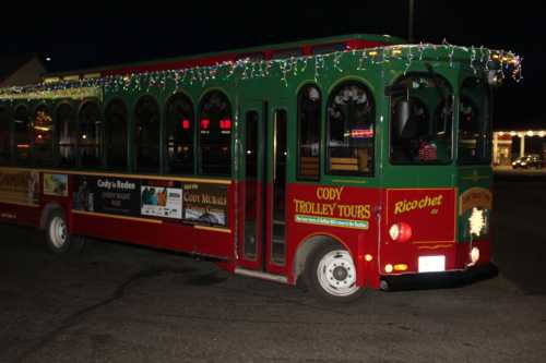 A festive green and red trolley bus decorated with lights, parked at night in a city setting.