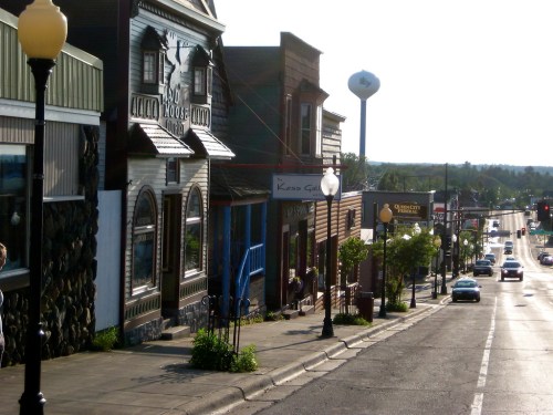 A scenic view of a small town street lined with shops, trees, and a distant water tower under a clear sky.