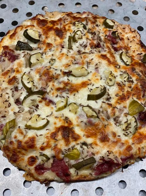 A freshly baked pizza topped with cheese, pickles, and herbs on a perforated baking tray.