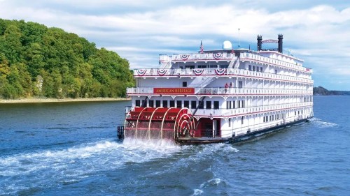 A large riverboat, the American Heritage, cruises on a calm river with lush green hills in the background.