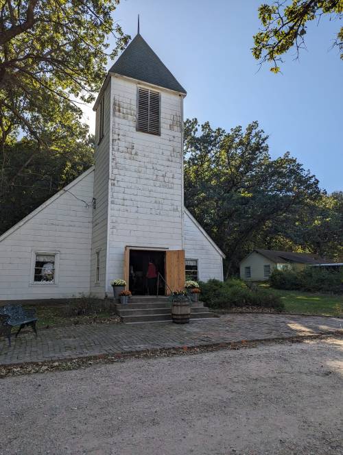 A rustic white church with a tall steeple, surrounded by trees and a gravel path, featuring a wooden door and flower pots.
