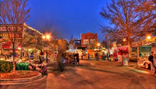 A festive outdoor market at dusk, with food trucks and twinkling lights, surrounded by trees and people enjoying the scene.