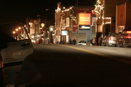 A festive street at night, lined with shops and illuminated by holiday lights, featuring a Wells Fargo sign.