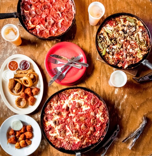 A table with two pizzas, a plate of fried food, and drinks, all set on a wooden surface.