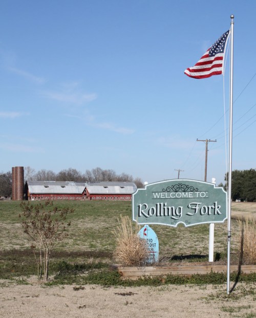 A welcome sign for Rolling Fork, with a barn and American flag in the background under a clear blue sky.