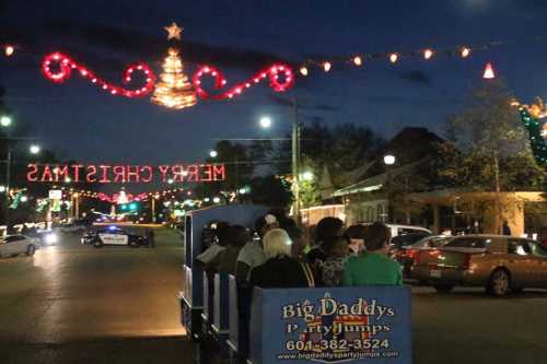 A festive night scene with a decorated street, Christmas lights, and a party vehicle filled with people.