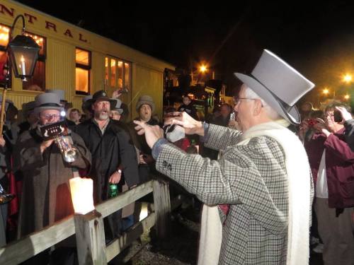 A crowd gathers at night, with a man in a top hat conducting musicians playing brass instruments.