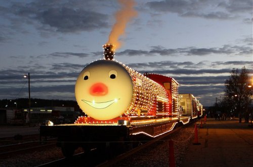 A brightly lit train with a smiling face and a red nose, decorated with lights against a twilight sky.