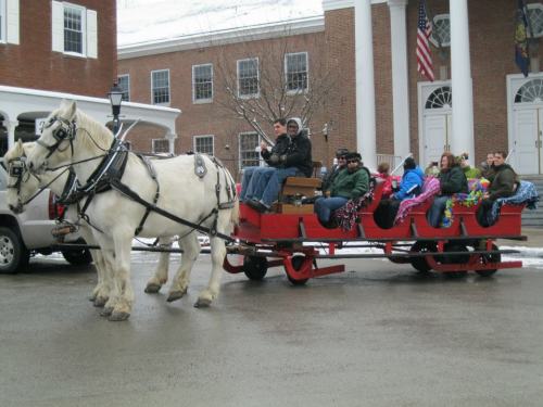 A horse-drawn sleigh carries passengers wrapped in blankets, parked in front of a brick building with flags.