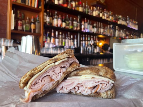 A close-up of a sliced sandwich with turkey and cheese on toasted bread, set against a blurred bar background.