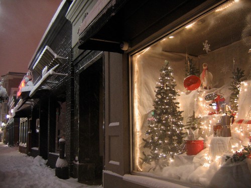 A snowy street scene featuring a festive shop window decorated with a Christmas tree and holiday lights.