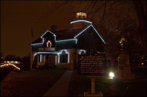 A historic lighthouse with festive lights at night, featuring a sign detailing its history and significance.