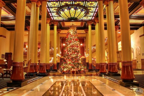 A beautifully decorated Christmas tree stands in a grand lobby with tall columns and a colorful stained glass ceiling.