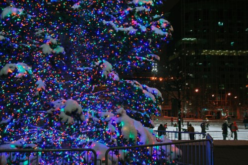 A brightly lit Christmas tree covered in snow, with colorful lights glowing against a dark night sky and ice skaters nearby.