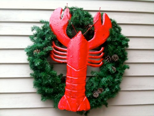 A festive wreath featuring a large red lobster, surrounded by green foliage and pinecones.