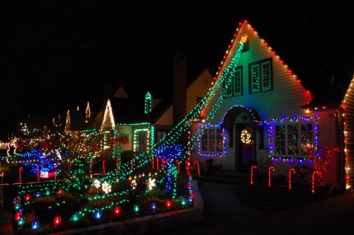 A cozy house decorated with colorful Christmas lights, surrounded by festive decorations in a nighttime setting.