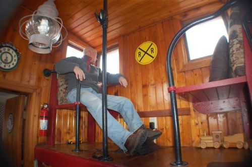 A person sits in a wooden room with a lantern, railroad sign, and toy train, surrounded by wooden walls and furniture.