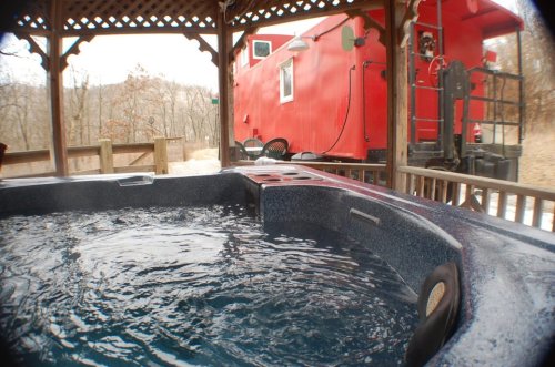 A hot tub under a wooden gazebo, with a red train car visible in the background and trees in the distance.