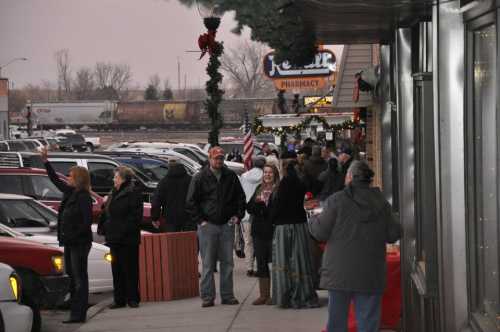 A busy street scene with people shopping, decorated storefronts, and cars parked along the road on a cloudy day.