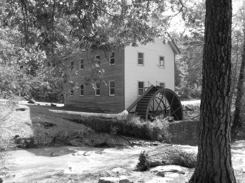 A historic watermill with a large wheel, surrounded by trees and a flowing stream, captured in black and white.