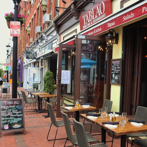Outdoor dining area of a restaurant with tables set, surrounded by shops on a brick-paved street.