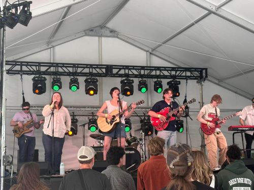 A band performs on stage under a tent, with musicians playing instruments and a crowd watching.