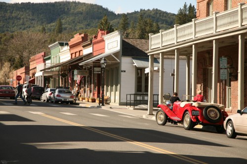 A historic street lined with colorful buildings and parked cars, featuring a vintage red car in the foreground.
