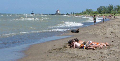 Three children lie on a sandy beach, playing in the sand, with waves and a lighthouse in the background.