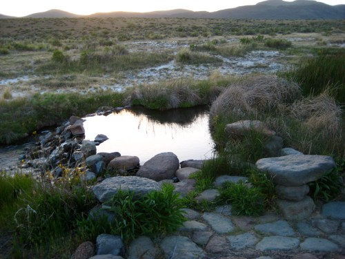 A serene natural pond surrounded by rocks and grass, with mountains in the background and a sunset glow.