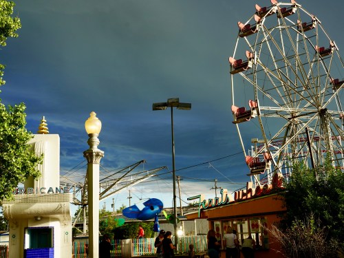 A carnival scene featuring a Ferris wheel, colorful booths, and a dramatic sky in the background.