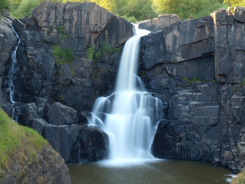 A serene waterfall cascading over dark rocks into a calm pool, surrounded by lush greenery.