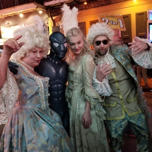 A group of four people in elaborate costumes, including a superhero, posing together on a festive street.