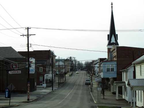 A quiet street scene in a small town, featuring a church steeple and lined with buildings and power lines.