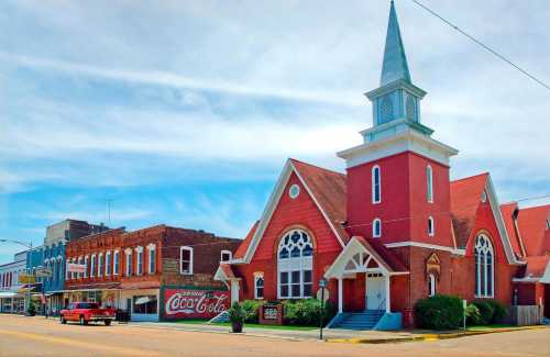 A red church with a steeple stands beside historic brick buildings and a vintage Coca-Cola mural in a small town.