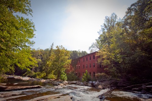 A red brick building nestled among trees by a river, with rocky banks and a clear blue sky above.