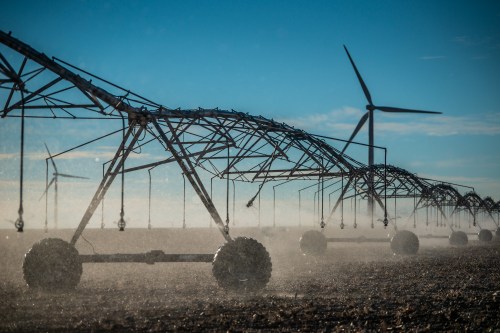 Irrigation system in a field with wind turbines in the background, dust rising under a clear blue sky.