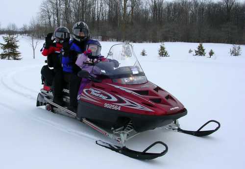 Three people wearing helmets ride a red snowmobile through a snowy landscape with trees in the background.