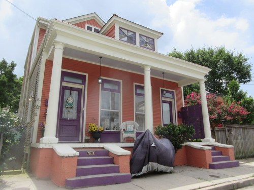 Colorful two-story house with purple accents, white columns, and a porch adorned with flowers and furniture.