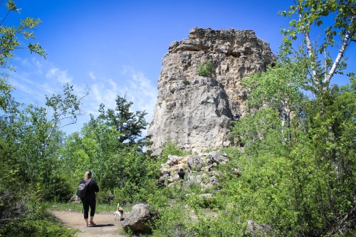 A person walks a dog on a trail near a large rock formation surrounded by greenery under a clear blue sky.