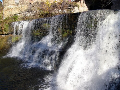A serene waterfall cascading over rocks into a calm river, surrounded by greenery and autumn foliage.